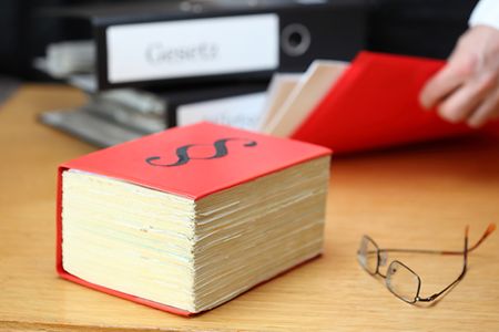 Rotes Gesetzbuch und Brille auf einem Tisch, im Hintergrund liegen Unterlagen und Aktenordner