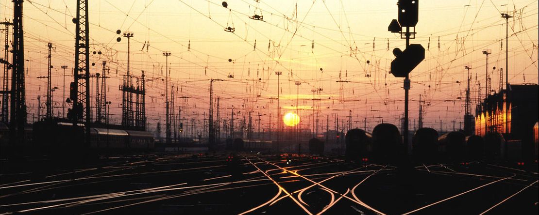 Bahnhofsvorfeld mit Gleisen und Oberleitungen bei Sonnenuntergang oder Sonnenaufgang.