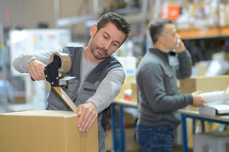 Mann verschickt ein Paket w&auml;hrend im Hintergrund ein zweiter Mann telefoniert.