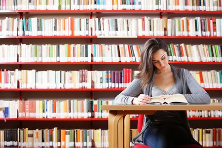 junge Frau sitzt in &ouml;ffentlicher Bibliothek an einem Tisch und liest in einem Buch