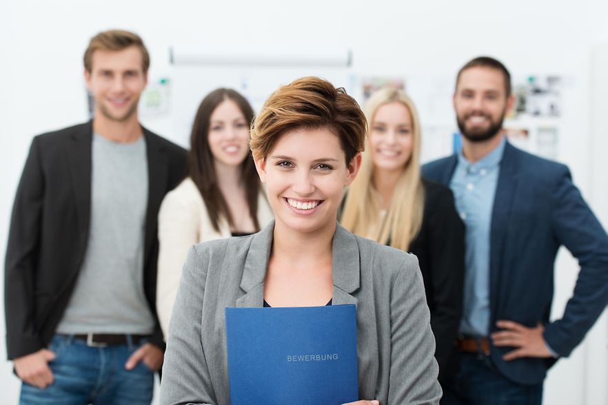Junge Frau h&auml;lt eine Bewerbungsmappe in der Hand. Im Hintergrund stehen vier weitere junge Menschen