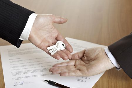Hand &uuml;bergibt einen Schl&uuml;ssel in eine andere Hand mit Vertrag im Hintergrund