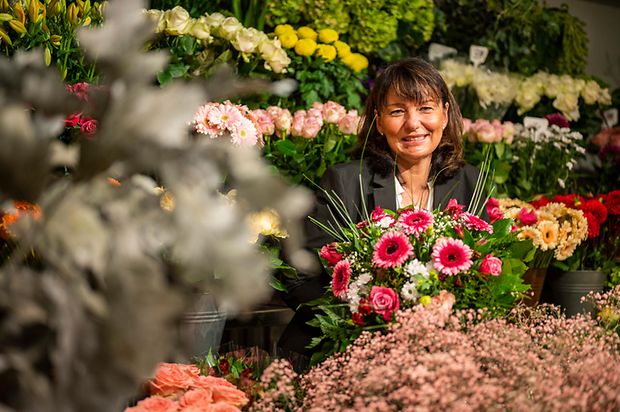 Eine Frau im schwarzen Jackett sitzt zwischen bunten Blumen.