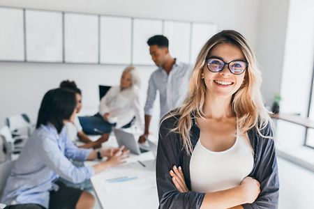 Eine blonde Frau mit Brille lehnt an einem Tisch, an dem mehrere Personen sitzen.