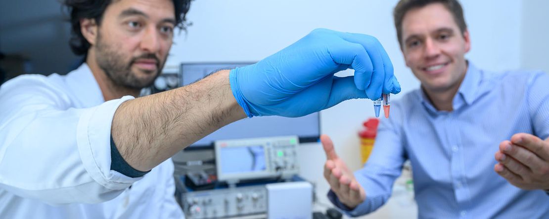 Zwei M&auml;nner stehen unter B&auml;umen vor einem Geb&auml;ude 
