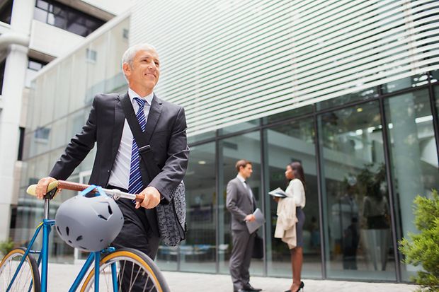 Ein Mann im Anzug schiebt ein Fahrrad an einem Unternehmensgeb&auml;ude vorbei. 
