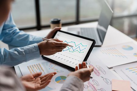 Symbolbild: 2 Business-Personen schauen auf ein Tablet mit Statistikkurven im Hintergrund Ausdrucke mit statistischen Daten