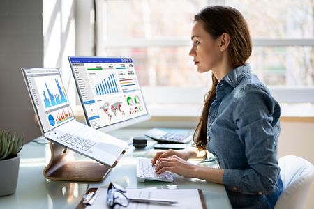 Symbolbild: Businessfrau am Schreibtisch sitzend betrachtet Ausdrucke mit statistischen Daten auf mehreren Monitoren