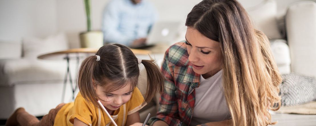 Mutter malt mit ihrem Kleinkind w&auml;hrend der Vater im Hintergrund im Homeoffice arbeitet