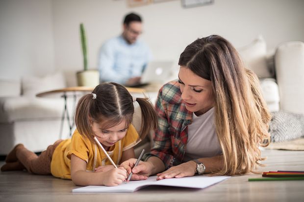 Mutter malt mit ihrem Kleinkind w&auml;hrend der Vater im Hintergrund im Homeoffice arbeitet
