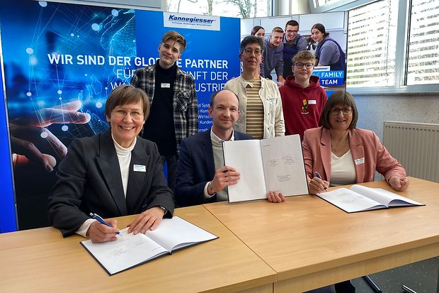 Vertreter von IHK, Unternehmen und Schule sitzen am Tisch und unterzeichnen die Vereinbarung, im Hintergrund stehen Jugendliche