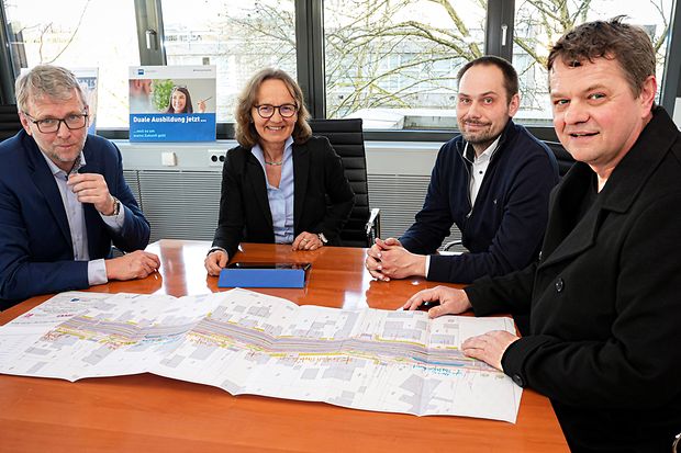 Vier Personen sitzen am Konferenztisch, vor ihnen liegt ein gro&szlig;er Umbauplan auf dem Tisch