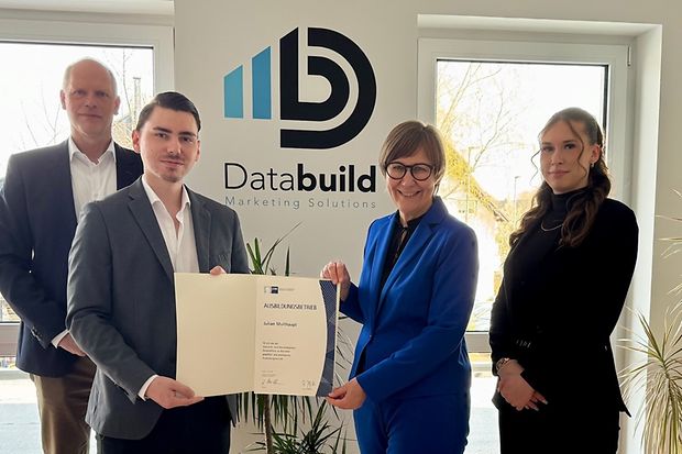Vier Personen vor dem Logo von "Databuild Marketing Solutions", eine Frau und ein Mann halten gemeinsam eine Ausbildungsurkunde.
