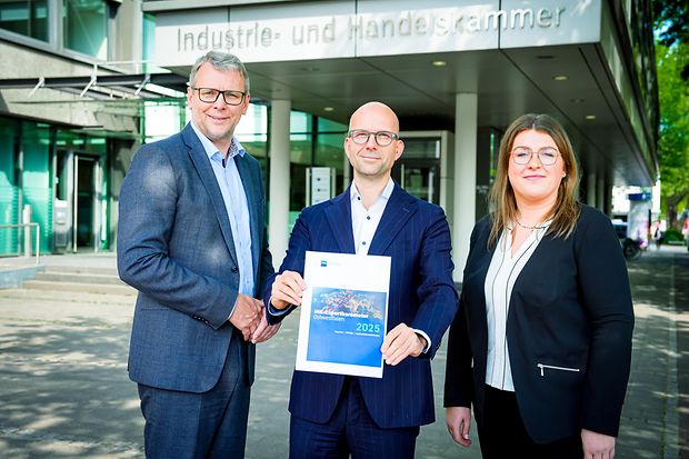 Drei Personen vor der IHK, eine Person h&auml;lt das IHK-Exportbarometer Ostwestfalen 2025.