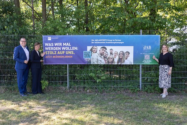 Drei Personen stehen vor einem Zaun mit Banner zur IHK-Ausbildungskampagne bei der JAB Anstoetz Group.