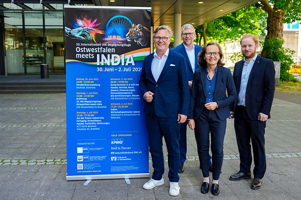 Vier Personen stehen neben einem Roll-Up mit dem Programm der &bdquo;20. IHK-Begegnungstage Ostwestfalen meets India&ldquo; in Bielefeld.