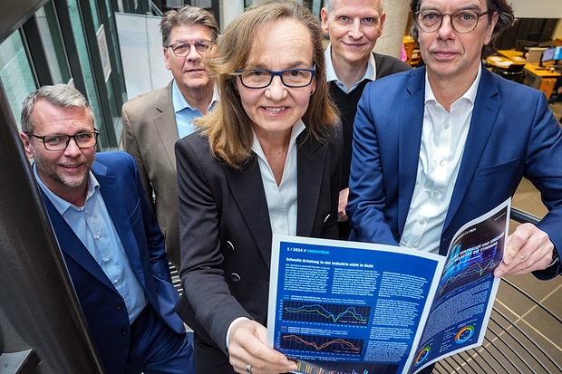 F&uuml;nf Personen in Businesskleidung stehen auf einer Treppe und halten einen ge&ouml;ffneten Konjunkturbericht in die Kamera.