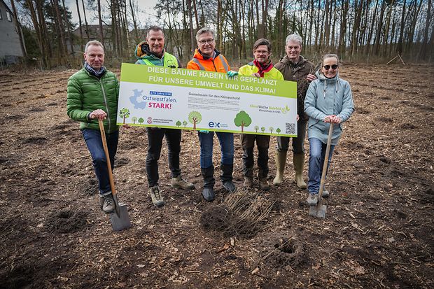 Sechs Personen mit Spaten halten ein Schild zur Baumpflanzaktion im Wald; Engagement f&uuml;r Umwelt und Klima.