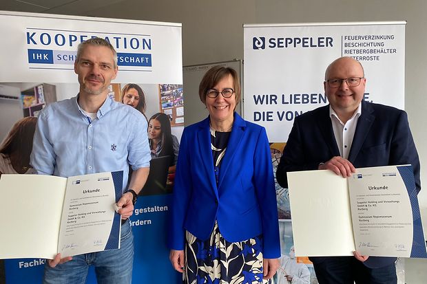 wei M&auml;nner und eine Frau halten feierlich unterzeichnete Urkunden vor IHK-Roll-ups