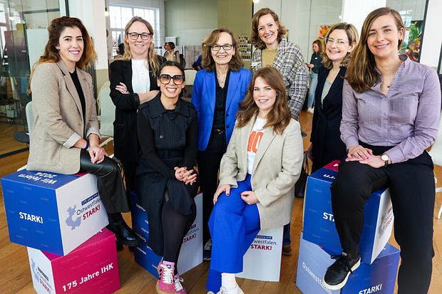 Acht Frauen, teils sitzend auf bunten W&uuml;rfeln mit IHK-Logo, posieren gut gelaunt in einem modernen Raum