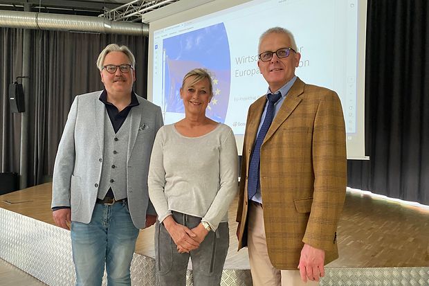 Drei Personen stehen vor einer Leinwand mit EU-Thema auf einer B&uuml;hne &ndash; Fokus auf wirtschaftliche Bildung.