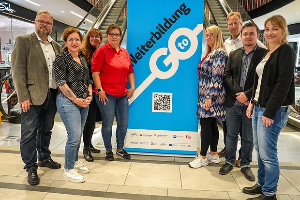 Zehn Personen stehen im Einkaufszentrum neben einem gro&szlig;en blauen Roll-up zur Aktion &bdquo;Weiterbildung to go&ldquo;.