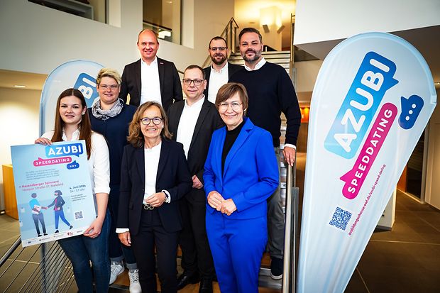 Gruppe von neun Personen steht auf einer Treppe mit Plakaten und Bannern zum &bdquo;Azubi-Speed-Dating&ldquo;.