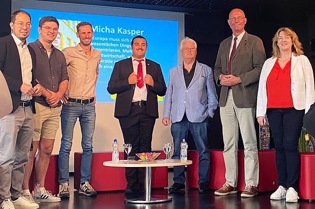 Sieben Personen stehen auf einer B&uuml;hne vor einer Pr&auml;sentation &ndash; politische Diskussion im Rahmen eines EU-Formats.