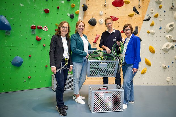 Vier Personen &uuml;bergeben Klettergurte vor einer bunten Kletterwand
