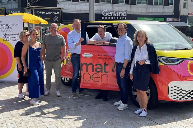 Gruppe von sieben Personen steht vor einem bunt folierten &bdquo;heimat shoppen&ldquo;-Auto