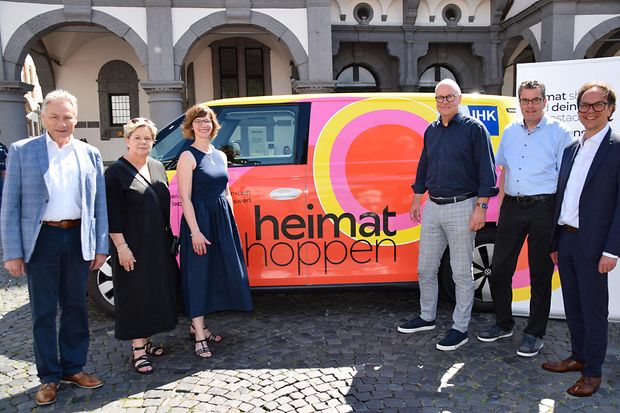 Sechs Personen posieren vor einem bunten Auto mit &bdquo;heimat shoppen&ldquo;-Aufdruck &ndash; Aktion im Stadtraum mit st&auml;dtischem Hintergrund.