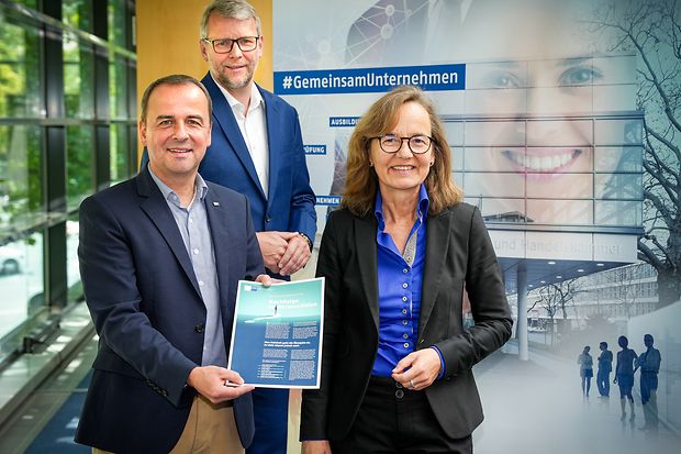 Zwei M&auml;nner und eine Frau stehen in Gesch&auml;ftskleidung mit einem Infoposter vor einem IHK-Banner