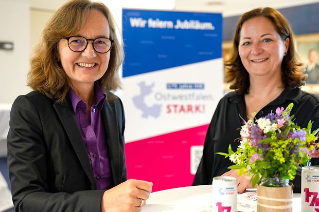 Zwei Frauen bei einer IHK-Veranstaltung in Ostwestfalen, im Vordergrund ein Stehtisch mit Blumen und Jubil&auml;ums-Getr&auml;nkedosen.