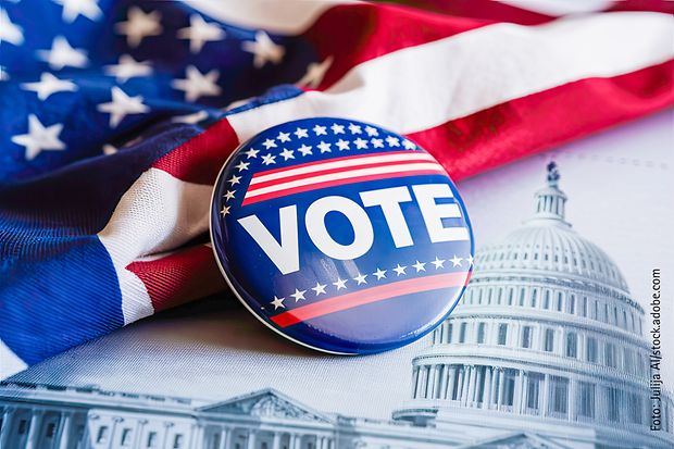 Wahl-Button mit Aufschrift &bdquo;VOTE&ldquo; auf US-Flagge und Kapitol-Hintergrund.