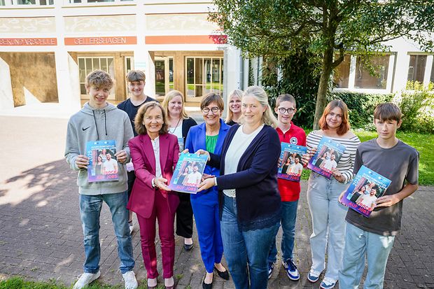 Gruppe Jugendlicher und Erwachsener steht vor Schule und h&auml;lt Ausbildungsatlas mit Aufschrift &bdquo;Bock auf Jobs&ldquo; in den H&auml;nden.