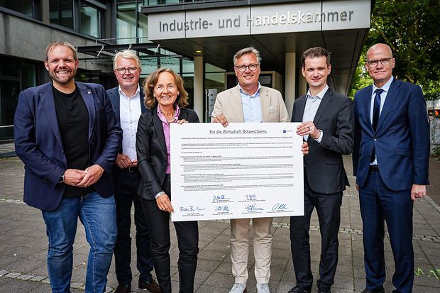 Sechs Personen stehen vor dem IHK-Geb&auml;ude in Bielefeld und halten ein gro&szlig;es Dokument zur Wirtschaft in Ostwestfalen.