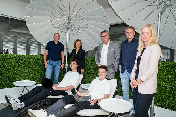 Gruppe unter Sonnenschirmen mit Laptops: Junge M&auml;nner mit Laptops auf Liegen, begleitet von Erwachsenen.