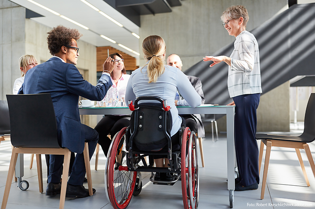 Symbolbild Inklusion: Diverse Gruppe im Business-Meeting, eine Frau im Rollstuhl sitzt am Konferenztisch.