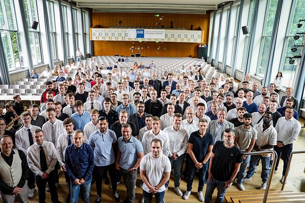 Eine gro&szlig;e Gruppe junger M&auml;nner und einige Frauen stehen in einer Aula zur Abschlussfeier