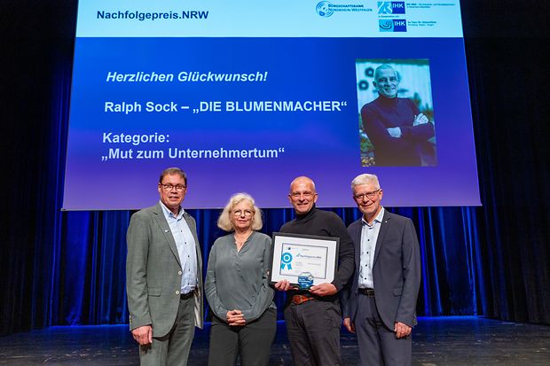 Vier Personen stehen auf einer B&uuml;hne, eine Person h&auml;lt eine Urkunde. Im Hintergrund ist Gl&uuml;ckwunsch auf dem Bildschirm zu sehen.