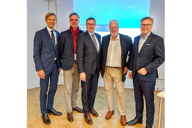 F&uuml;nf M&auml;nner im Anzug bei einer Veranstaltung, stehend vor einer Pr&auml;sentationsfolie.