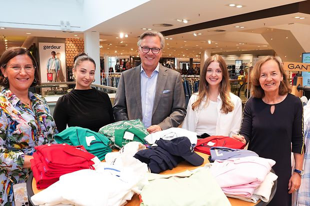 F&uuml;nf Personen stehen in einem Modegesch&auml;ft an einem Tisch mit zusammengefalteter Kleidung. Alle l&auml;cheln in die Kamera.