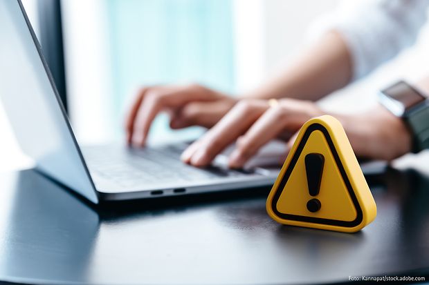 H&auml;nde tippen auf Laptop-Tastatur, im Vordergrund steht ein gelbes Warnsymbol mit Ausrufezeichen &ndash; Hinweis auf digitale Gefahr.