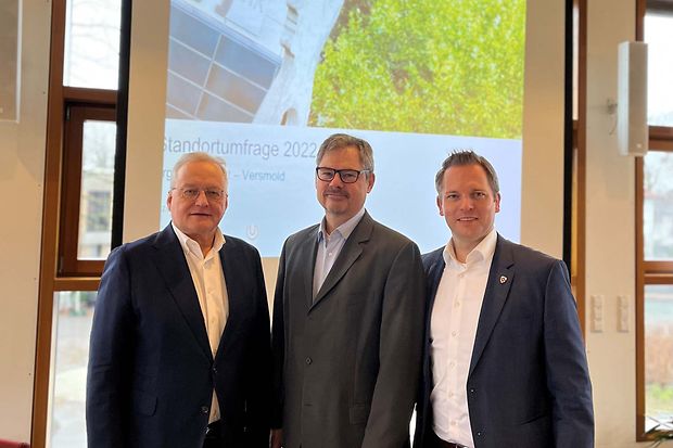 Drei M&auml;nner in Anz&uuml;gen stehen vor einer Leinwand mit einer Pr&auml;sentation zur Standortumfrage 2022.