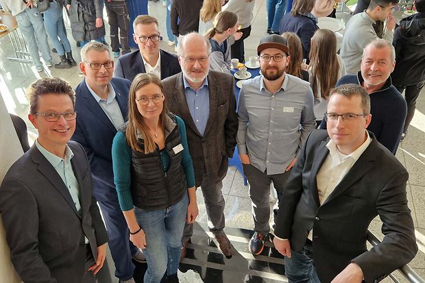 Gruppe von acht Personen, die l&auml;chelnd in die Kamera schauen, bei einer Auftaktveranstaltung in einem hellen Foyer.
