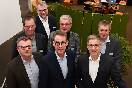 Sieben M&auml;nner in Businesskleidung stehen in einem modernen B&uuml;rogeb&auml;ude auf einer Treppe und blicken in die Kamera.