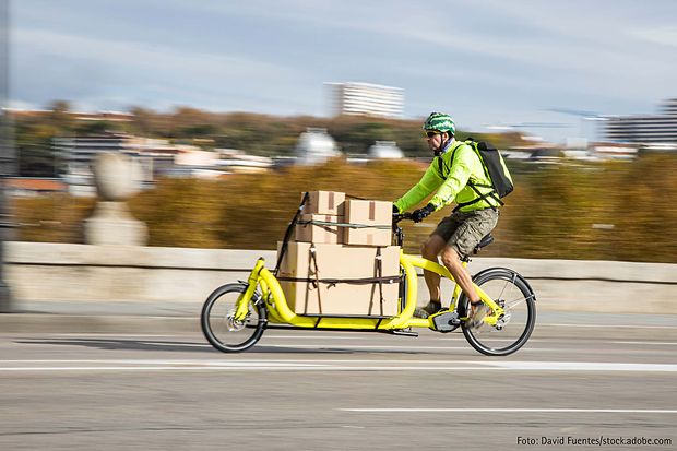 Mann f&auml;hrt mit gelbem Lastenrad, beladen mit Kartons, &uuml;ber eine Stra&szlig;e
