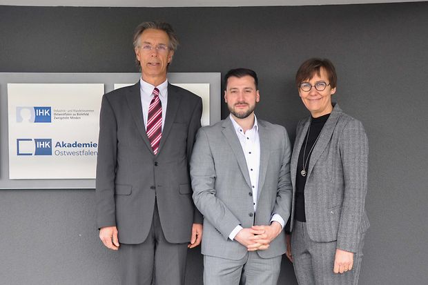 Drei Personen in grauen Anz&uuml;gen stehen vor einem Schild der IHK Akademie Ostwestfalen in Minden.
