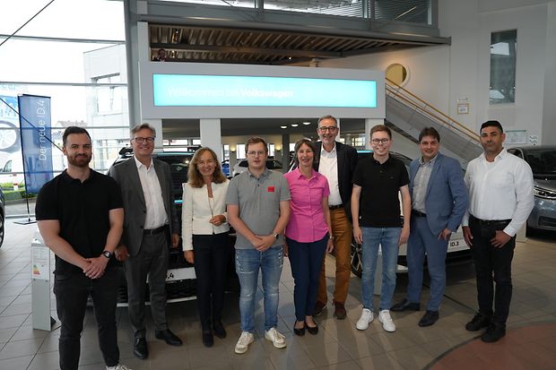 Neun Personen stehen in einer Autohalle vor einem Schild &bdquo;Willkommen bei Volkswagen&ldquo;.