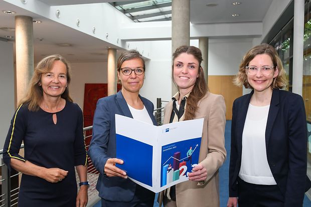 Vier Frauen im Business-Outfit halten gemeinsam eine Mappe mit dem Titel &bdquo;Gr&uuml;ndungsreport&ldquo;.